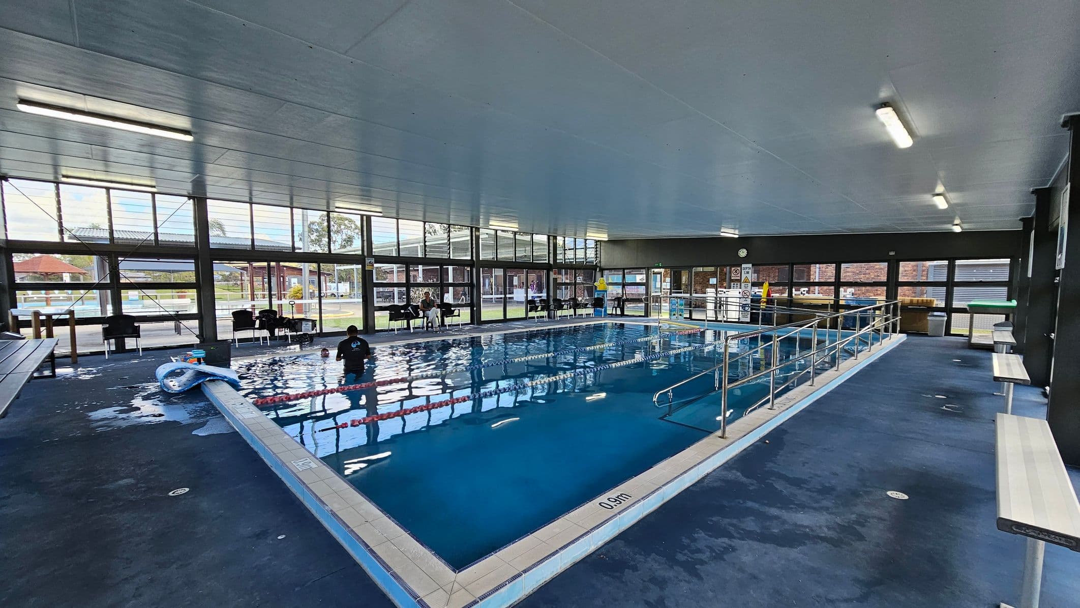 Indoor swimming pool with a blue-tiled floor and metal railings on the right. A person is in the water near the pool's edge. On the left, pool equipment, including a float, is visible. The background features large windows with views of an outdoor area, including a red-roofed structure and greenery. The ceiling is grey with fluorescent lights, and there are benches along the right side.