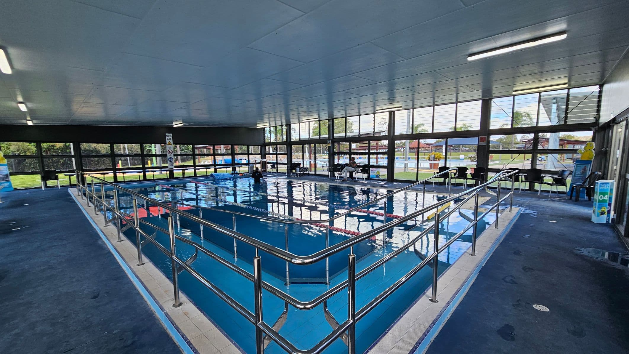 Indoor swimming pool with a ramp and metal railings leading into the water. The pool is surrounded by a blue-tiled floor. Large windows line the walls, allowing natural light to enter and providing a view of the grassy outdoor area. Several chairs are placed along the windows, and there are signs and a small bin near the entrance. The ceiling is fitted with fluorescent lights.