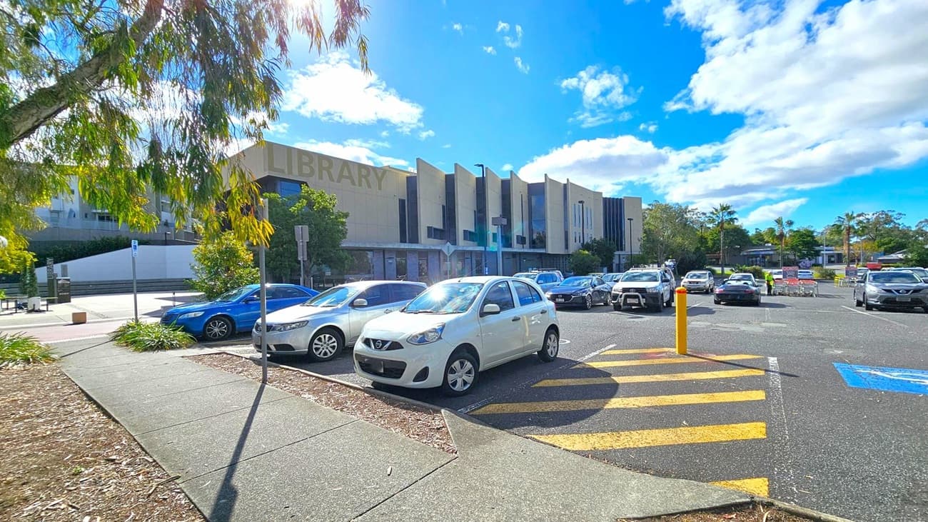 A car park with several vehicles, including a white car in the foreground. On the left, a tree with green leaves partially shades the area. The background features a large building with the word "LIBRARY" visible on its facade. The sky is blue with scattered clouds. A yellow-striped pedestrian crossing and a blue disabled parking space are visible on the asphalt.