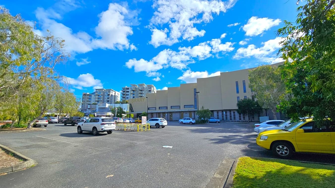 A car park with several vehicles, including a prominent yellow car on the right. In the background, a large beige building with the word "LIBRARY" is visible. To the left, there are trees with sparse foliage, and several multi-storey buildings can be seen in the distance. The ground is paved, and there is a small patch of grass near the yellow car.