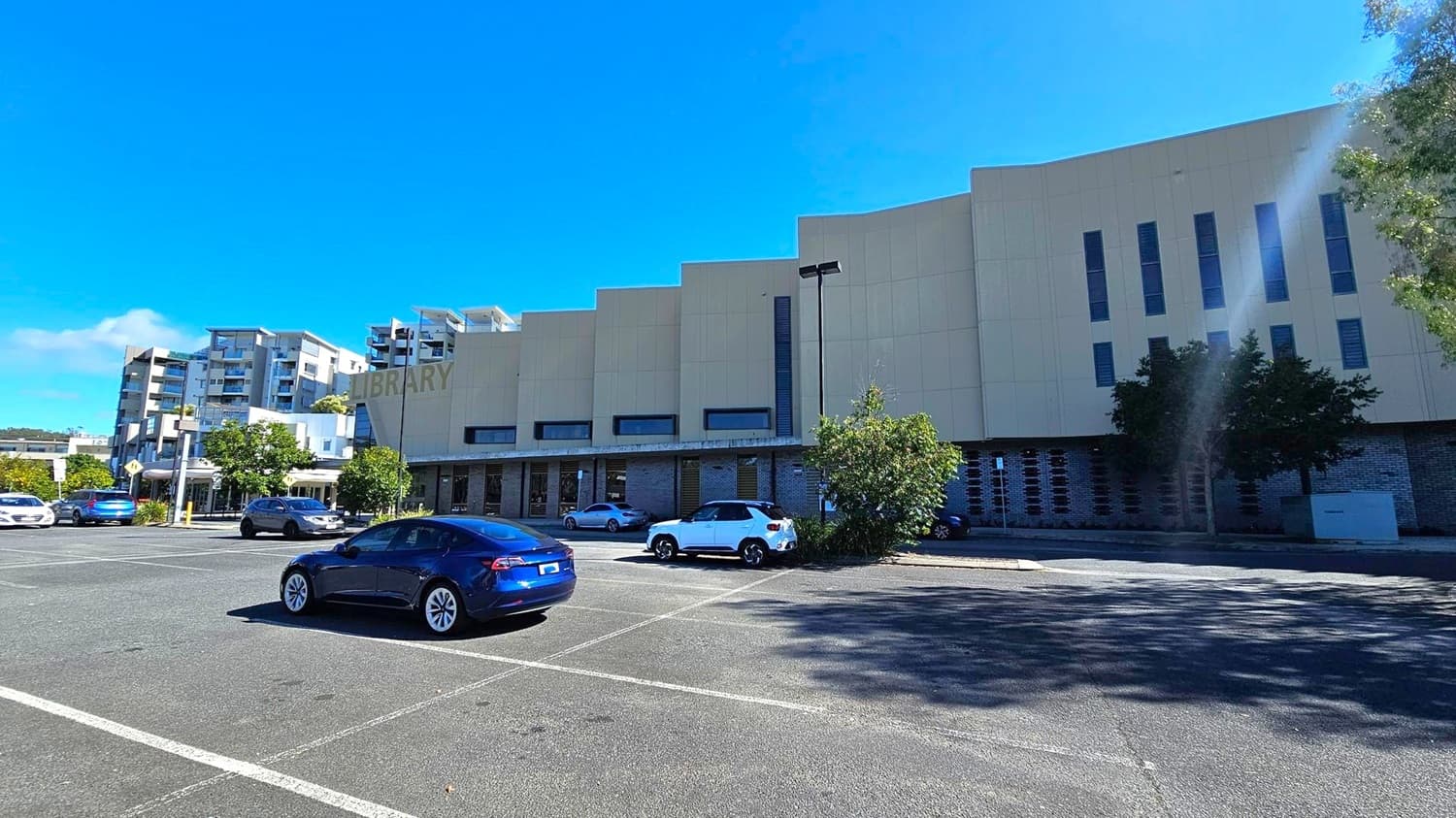 A large, modern building with the word "LIBRARY" on its facade is on the right. The structure has a series of vertical windows and a light-coloured exterior. In the foreground, a car park with several vehicles, including a blue car and a white SUV. On the left, there are multi-storey residential buildings with balconies. Trees and shrubs are scattered around the area, providing some greenery. The ground is paved with marked parking spaces.