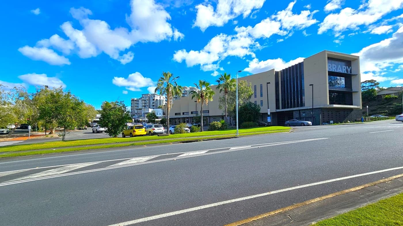 A modern building with the word "BRARY" visible on the right side, likely part of a library sign. The structure is surrounded by palm trees and other greenery. Several cars are parked in a lot to the left, including a yellow one. The foreground features a multi-lane road with white markings. In the background, there are additional buildings and more trees under a blue sky with scattered clouds.