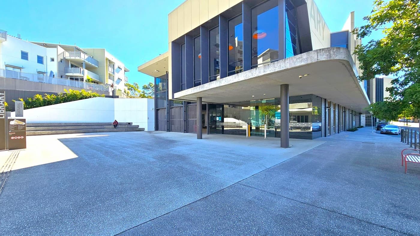 On the left, there is a modern building with white walls and balconies, surrounded by greenery. A person is sitting on steps near a sign that reads "Library" and "Cultural". In the centre, a large building with tall glass windows and a protruding roof supported by pillars. On the right, a tree provides shade over a parked car and a red bench. The ground is paved with a textured surface, and the area is well-lit by natural light.