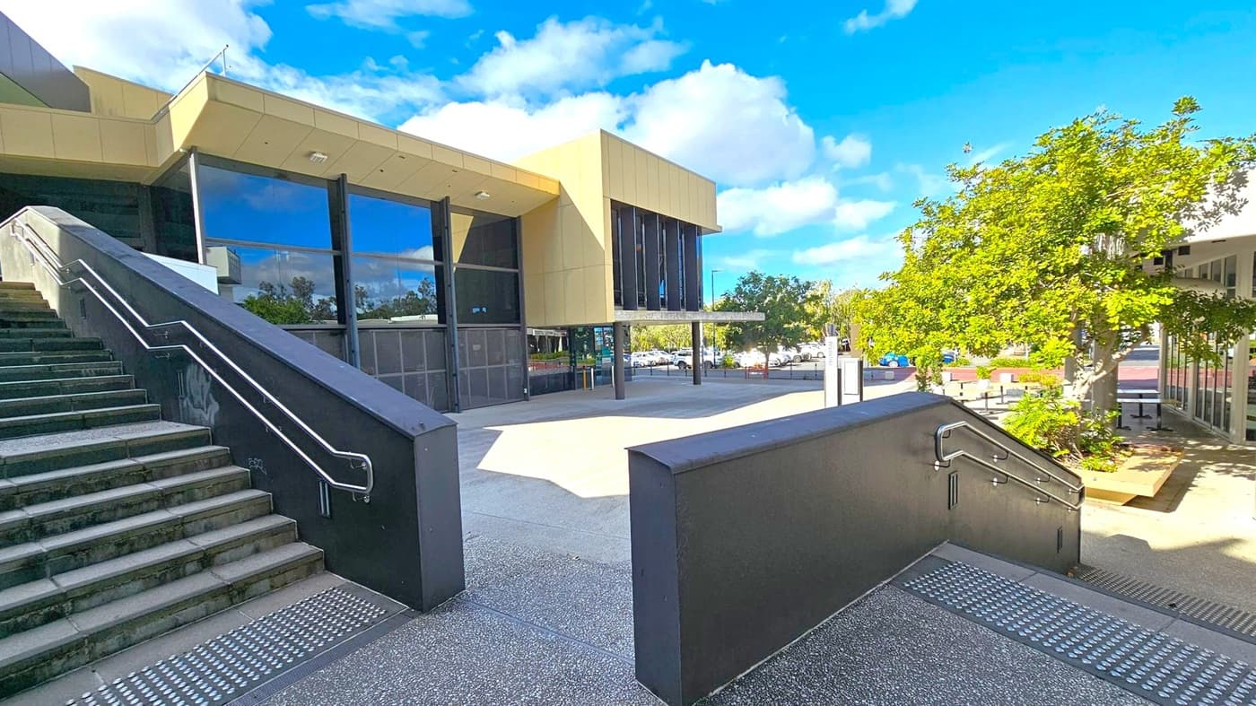 A modern building with large glass windows and beige cladding is in the centre. On the left, a wide staircase with metal railings leads up to the entrance. On the right, a tree with green foliage provides shade over a paved area. In the background, a car park is visible with several parked cars. The ground is a mix of textured and smooth concrete surfaces.