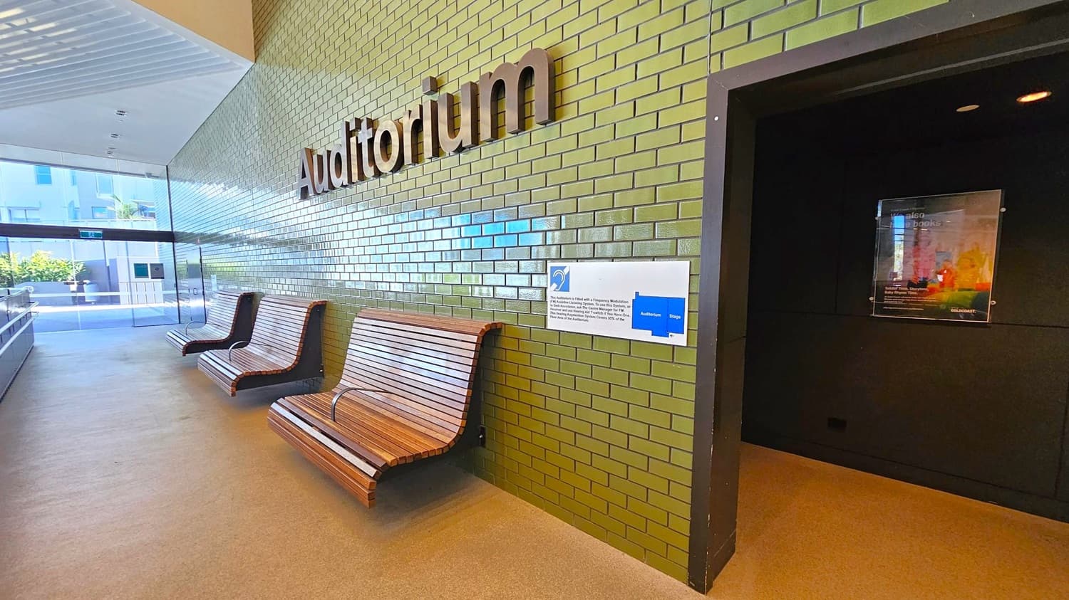 A hallway with green tiled walls and a sign reading "Auditorium" in large letters. On the left, there are three wooden benches with curved designs. A glass door at the end of the hallway reveals an outdoor area with plants. On the right, a dark entrance leads into another room, with a poster displayed on the wall. The floor is a light-coloured, smooth surface, and the ceiling is white with recessed lighting.