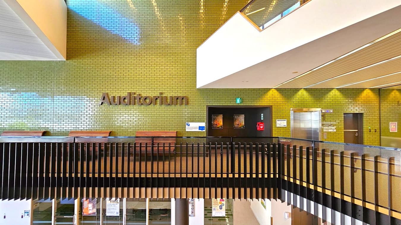 A modern interior with a green-tiled wall featuring the word "Auditorium" in large letters. Below, there are wooden benches and a black railing. On the right, a lift with metallic doors and a dark doorway are visible. The ceiling has wooden panels with embedded lights. The floor is not visible, but the lower level can be seen through the railing, showing glass doors and posters.