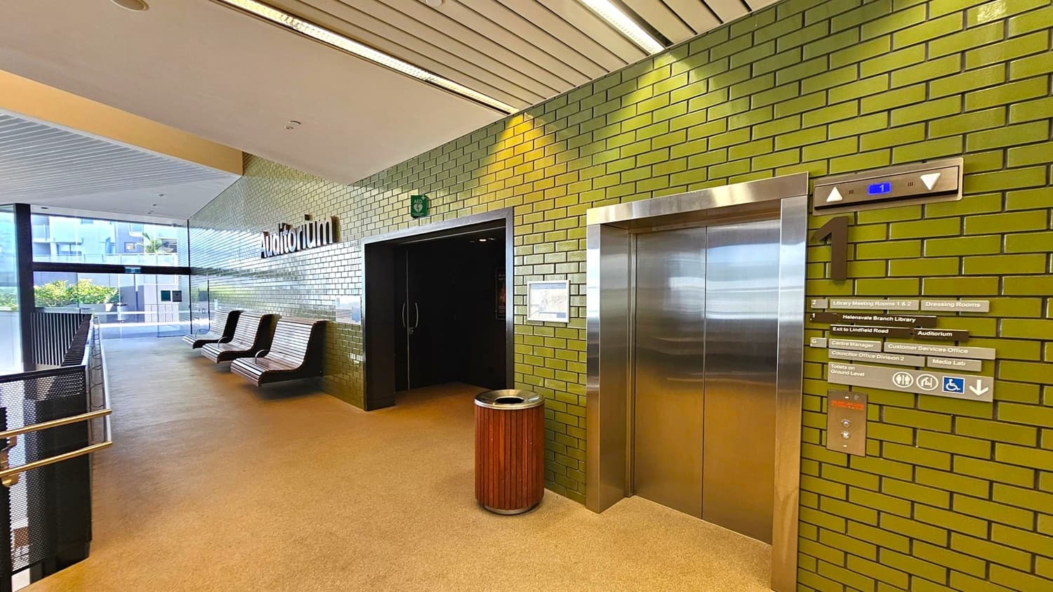 A hallway with green brick walls and a beige carpeted floor. On the right, there's a stainless steel lift with floor indicators and directional signs. A wooden rubbish bin is placed nearby. The left side features a row of modern, curved benches against the wall, leading to a glass window with a view of greenery outside. The ceiling is lined with white panels and fluorescent lights. An entrance to an auditorium is visible next to the lift.