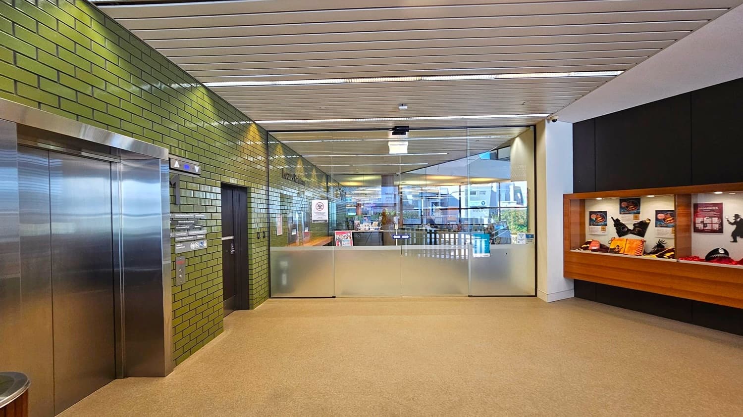 A hallway with a green tiled wall on the left featuring a stainless steel lift and a black door. The ceiling has a slatted design. In the centre, there's a glass partition with a frosted lower section, leading to another area. On the right, a display case with various items and posters is mounted on a wooden panel. The floor is carpeted in a light brown colour.
