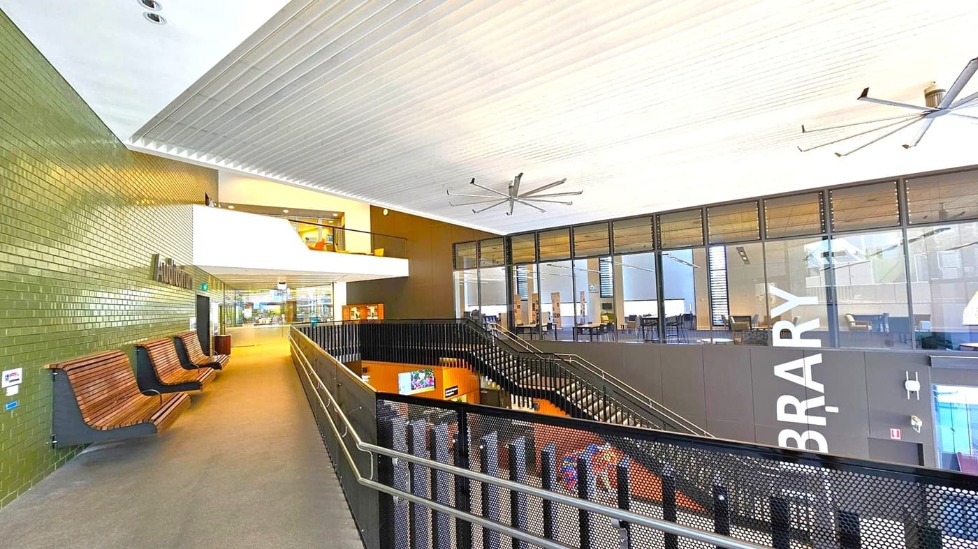 A modern interior with a high ceiling featuring large fans. On the left, there's a green-tiled wall with wooden benches and a sign for an auditorium. The right side has large glass windows with a view into a library area. A black metal railing lines the walkway, leading to a staircase descending to a lower level. The floor is carpeted, and the space is well-lit with natural and artificial light.