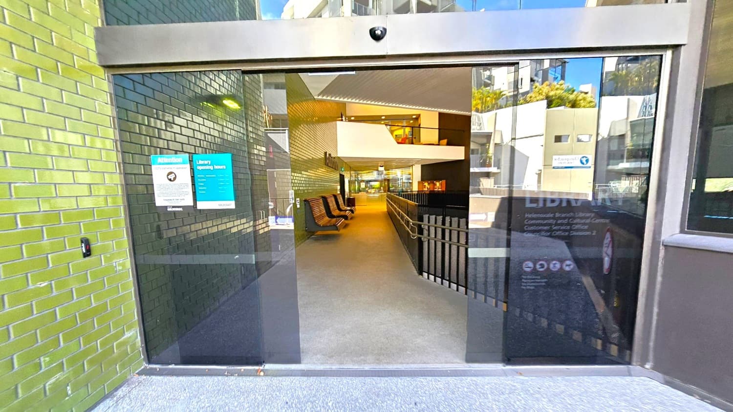 Glass entrance doors with signs about CCTV and library opening hours. The left wall is covered in green tiles. Inside, a corridor with wooden benches on the left and a railing on the right leads to a well-lit interior space. The floor is carpeted, and the ceiling has a modern design with recessed lighting. Reflections of nearby buildings are visible on the glass.
