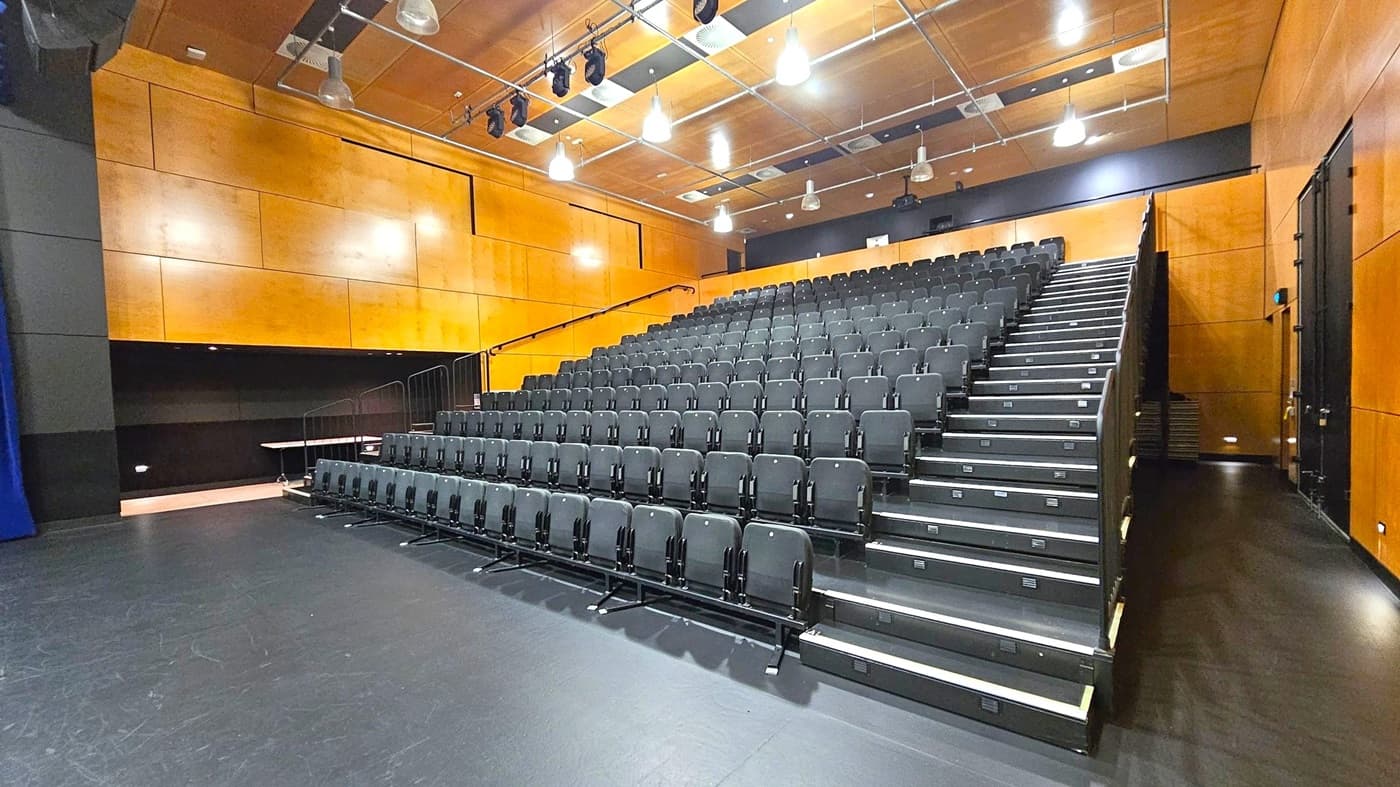 A theatre with tiered seating on the right, featuring rows of black chairs. The floor is dark and smooth. The walls are clad in light wood panelling. The ceiling is also wooden, with exposed lighting fixtures and ventilation ducts. The left side has a small alcove with a table, and the right side has a narrow walkway. The space is well-lit with overhead lights.
