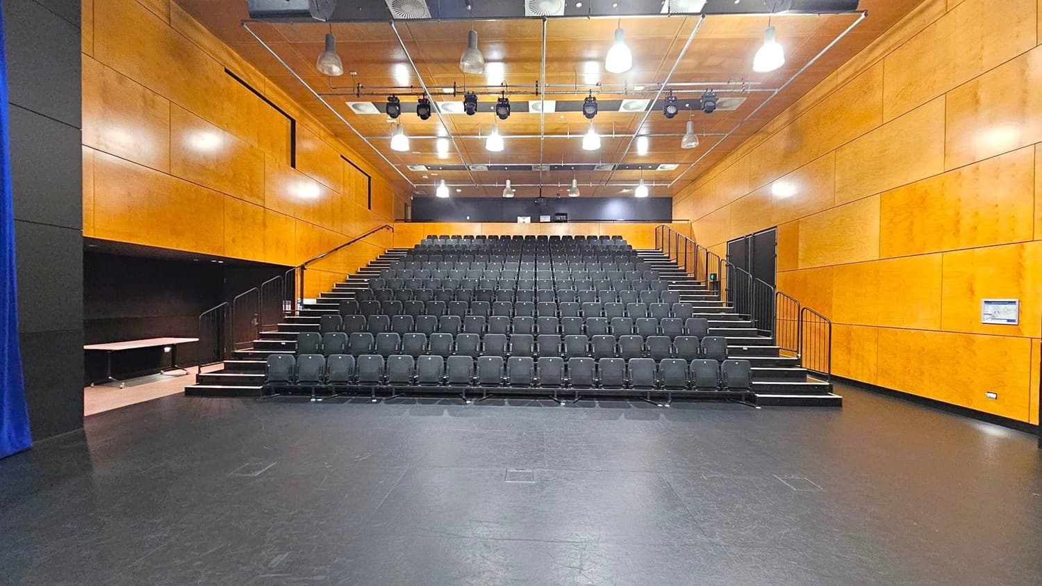 A theatre with tiered seating in the centre, featuring rows of black chairs. The walls are clad in light wood panelling. The ceiling is equipped with multiple hanging lights and stage equipment. On the left, a blue curtain is partially visible, while the right side has a wall-mounted sign. The floor is dark and smooth, contrasting with the light wood of the walls.