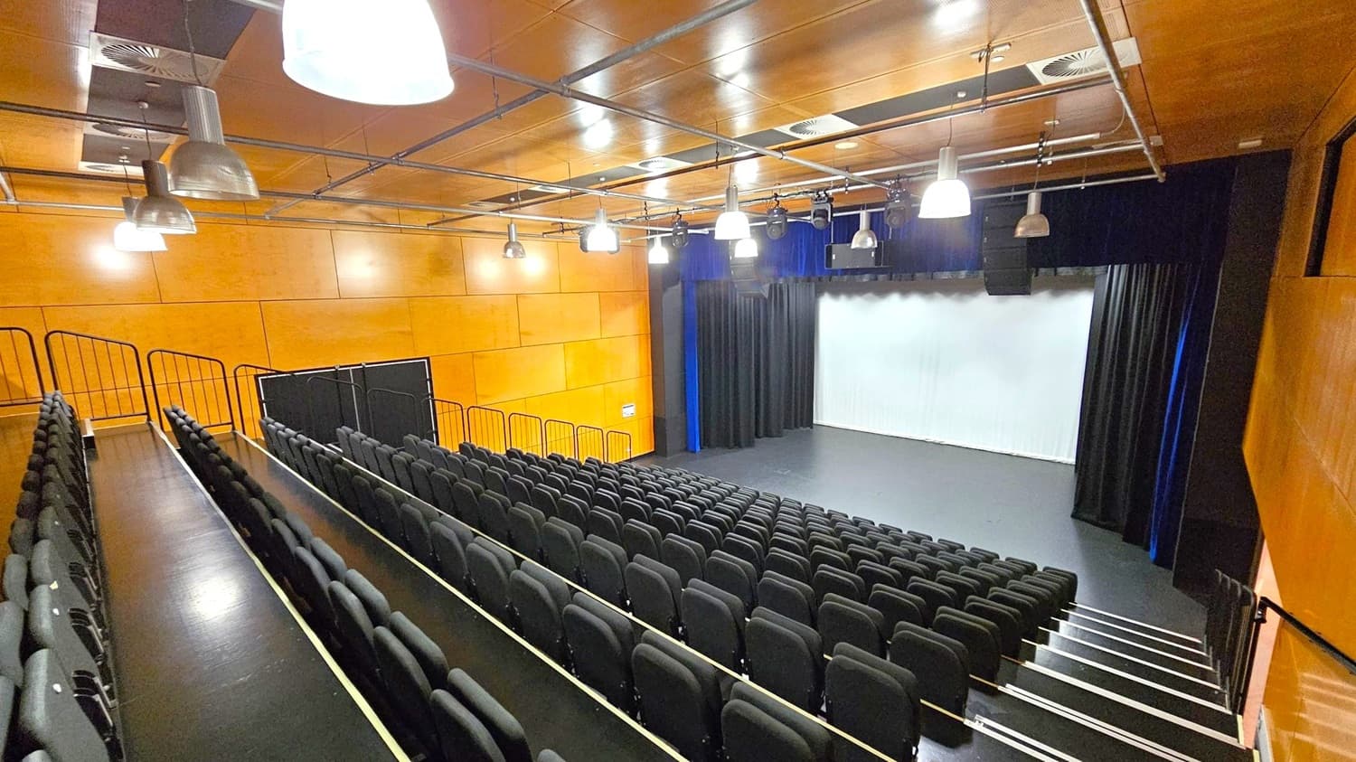 A modern auditorium with tiered seating on the left, featuring rows of black chairs. The stage is on the right, with a white screen and black curtains. The walls are wooden, and the ceiling is also wooden with exposed ductwork and several large, industrial-style pendant lights. The floor is dark, matching the seating area.