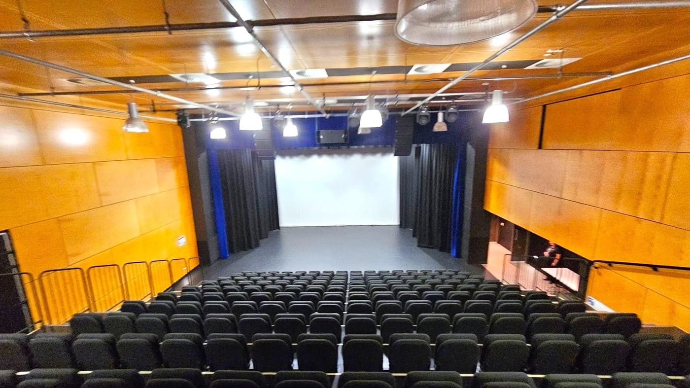A theatre with rows of black seats facing a stage. The stage has a white backdrop and is flanked by black curtains. The walls are covered in light wood panelling. The ceiling is also wooden with exposed metal beams and several hanging lights. On the right, there is a small control booth with a person seated inside. The floor is dark, matching the seats.