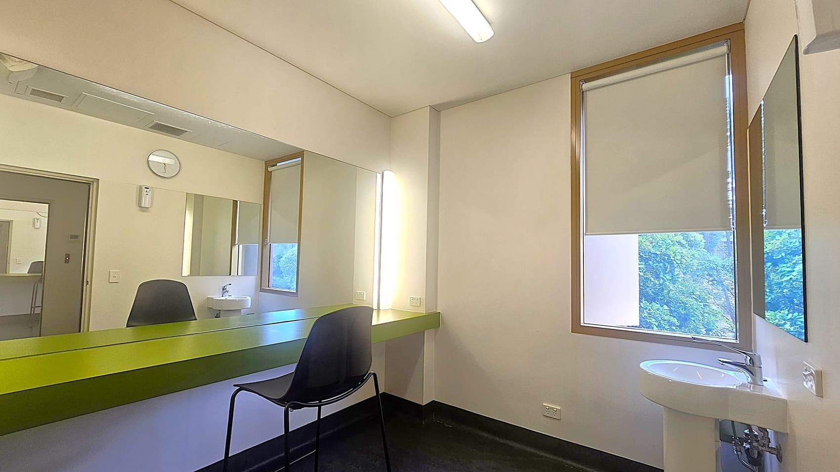 A room with a large mirror on the left wall, above a long green counter. A black chair is positioned in front of the counter. On the right, a window with a partially drawn blind reveals some greenery outside. Below the window is a small white sink. The ceiling has a fluorescent light fixture, and a clock is mounted on the wall above the mirror. The floor is dark, contrasting with the light-coloured walls.