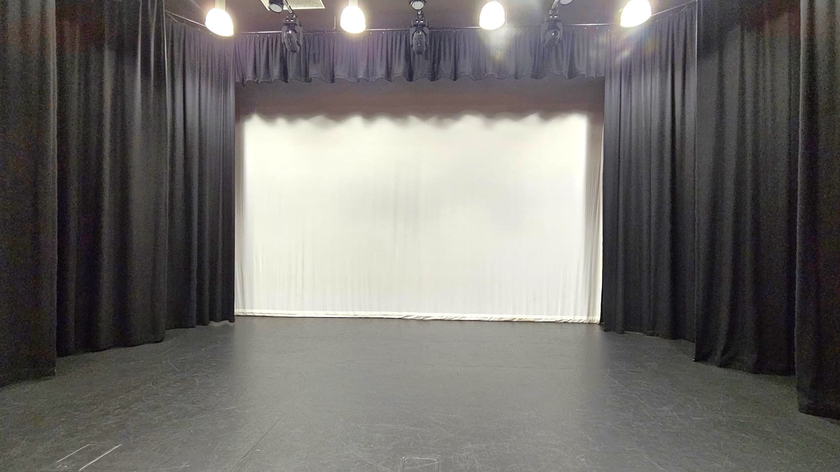 A stage with a black floor and black curtains on both sides. The background features a large white screen or backdrop. Overhead, several stage lights are mounted, casting light onto the stage area. The ceiling is dark, blending with the lighting equipment.