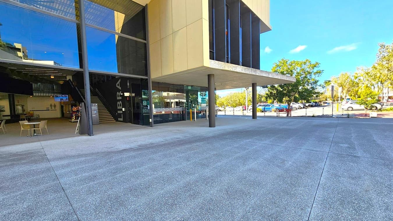 A modern building with large glass windows on the left, revealing an interior with tables, chairs, and a staircase. The word "LIBRA" is visible on the wall near the entrance. The building has a beige exterior with black columns supporting an overhang. On the right, there is a car park with several vehicles and trees in the background. The ground is paved with grey concrete.