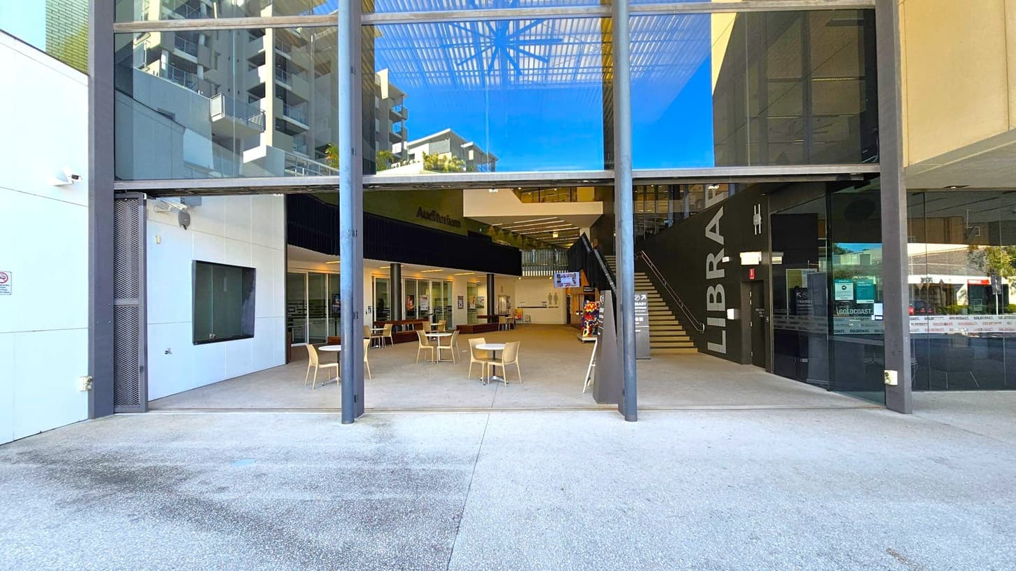 The image shows the entrance to a modern building with large glass windows. In the centre, there are tables and chairs arranged in an open area. To the right, a staircase leads upwards, with the word "LIBRARY" visible on the wall. The left side features a white wall with a dark window. The floor is a mix of concrete and tiled surfaces, and the ceiling has a grid-like structure with reflections of nearby buildings visible in the glass.