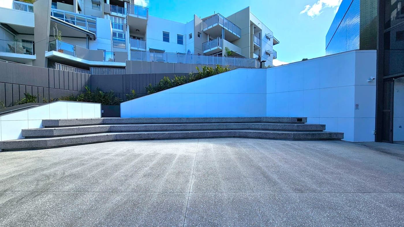 In the foreground, there is a wide, grey concrete area leading to a series of tiered steps. The steps are made of a darker material and are positioned centrally. In the background, modern apartment buildings with balconies and glass railings are visible. The left side features a dark panelled wall with some greenery at its base. The right side has a smooth, white wall with a slight curve. The sky is partially visible above the buildings.