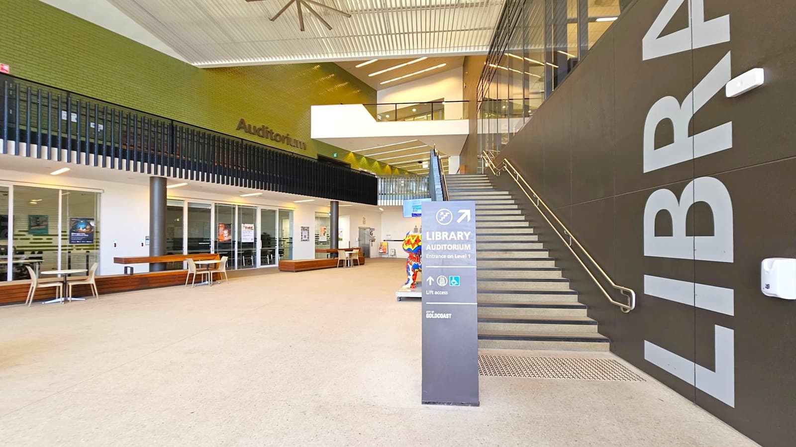 A modern interior space with a staircase on the right, featuring large letters spelling "LIBRARY" on the wall. A sign in front of the stairs indicates directions to the library and auditorium. On the left, there are glass doors with tables and chairs outside. Above, a green brick wall with the word "Auditorium" and a black railing. The ceiling has a large fan and linear lighting. The floor is light-coloured and smooth.