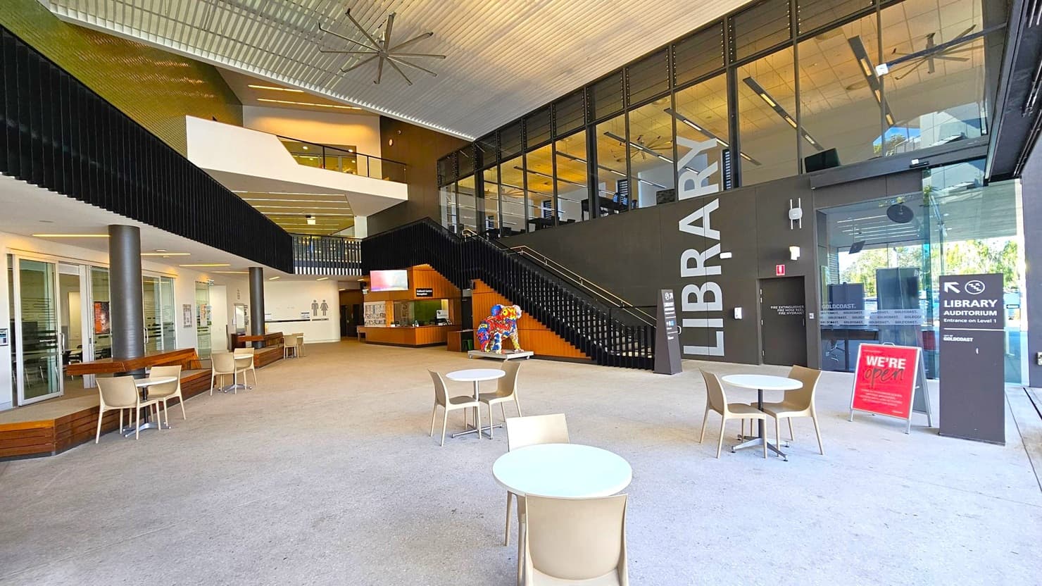 A spacious library entrance with a high ceiling and large fan. On the right, a black wall with "LIBRARY" in large letters and a glass entrance. A staircase leads to an upper level with glass panels. In the centre, a colourful sculpture is near the stairs. On the left, a seating area with tables and chairs, and a reception desk. The floor is light-coloured, and there are signs indicating the library is open.
