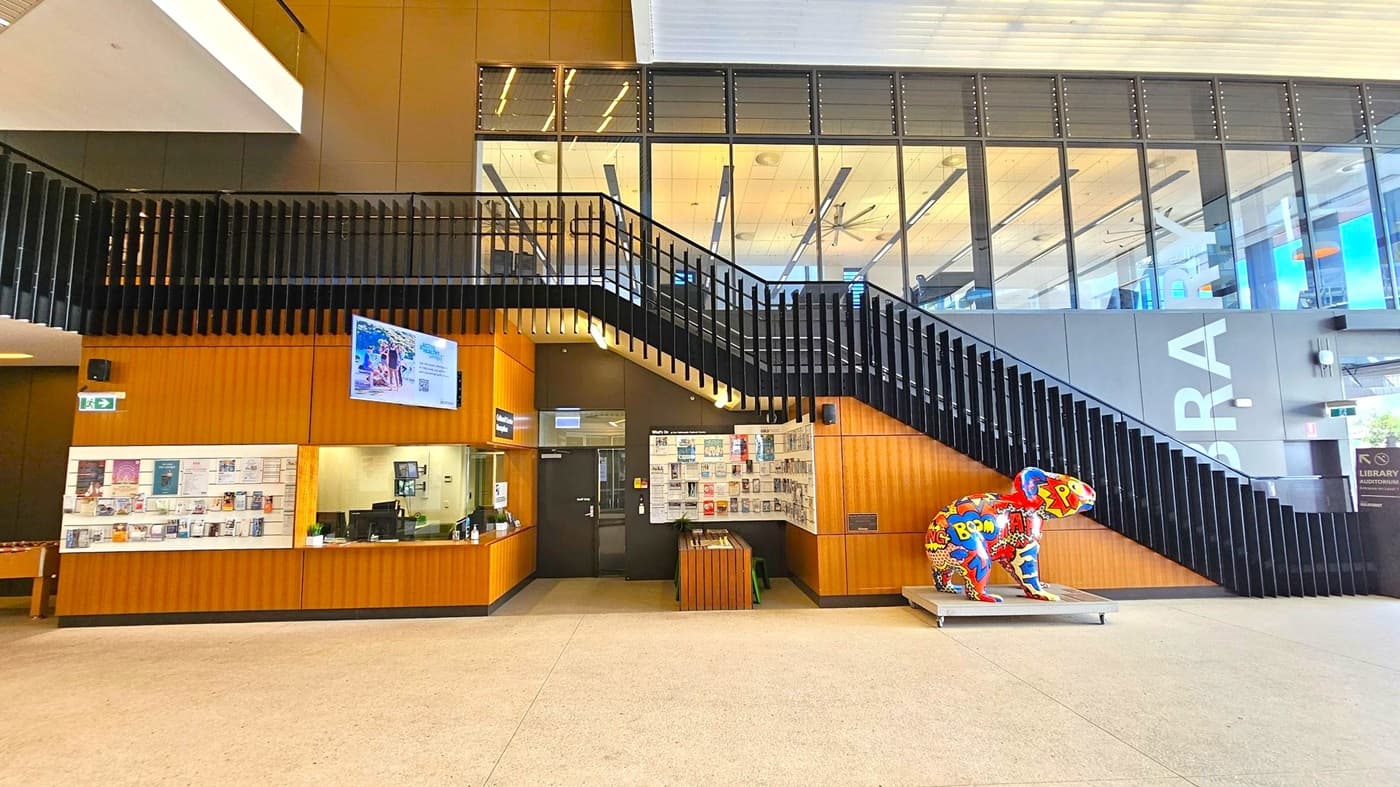 A modern interior with a staircase featuring black railings on the right. Below the stairs, a colourful sculpture of a koala with comic-style graphics is displayed. On the left, a wooden reception desk with a digital screen above it and a wall covered in posters and notices. The floor is light-coloured, and the ceiling has a grid pattern with visible lighting. Large windows above the staircase reveal an upper level with visible ceiling fans and lights.