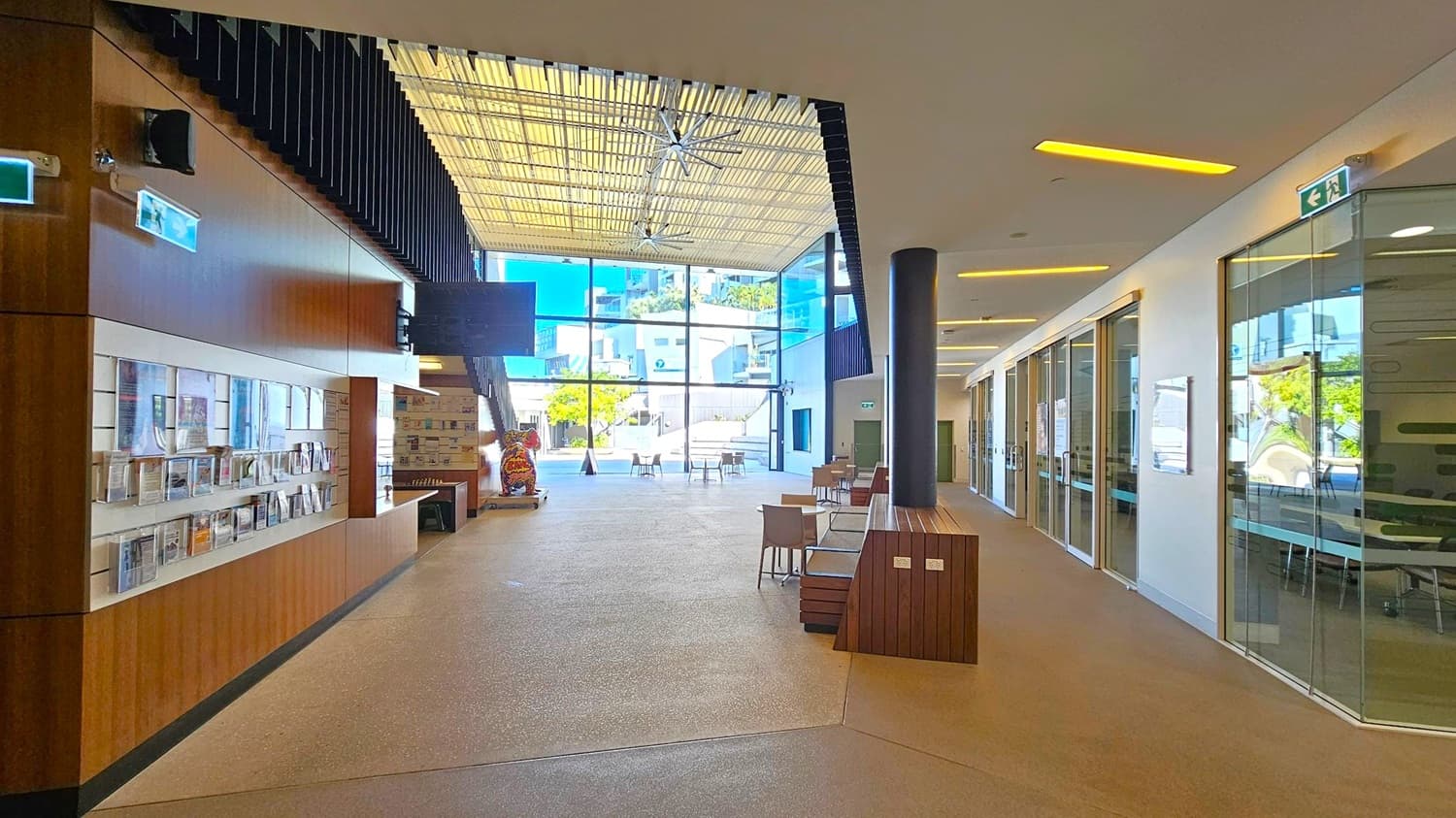 A spacious interior with a high ceiling featuring a large fan. On the left, there is a wooden wall with shelves displaying brochures and magazines. The right side has glass-walled rooms with visible tables and chairs inside. In the centre, there are tables and chairs arranged in an open area. The background shows large glass windows with a view of a courtyard and buildings outside. The floor is a light-coloured polished surface.