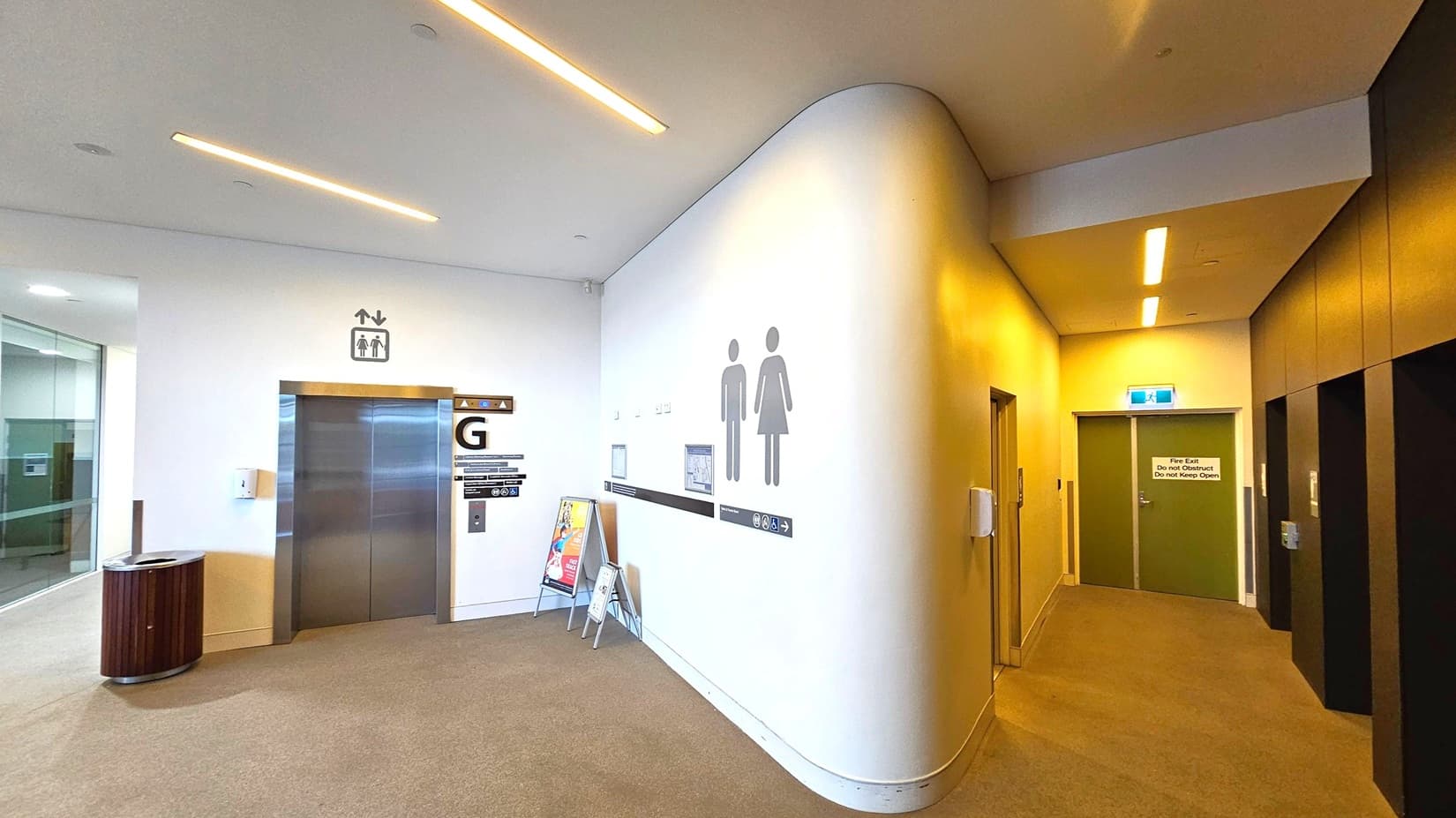 A corridor with a beige carpeted floor and a white ceiling featuring recessed lighting. On the left, there's a lift with a "G" sign above it, a bin nearby, and a glass wall. In the centre, a curved wall displays male and female restroom symbols. On the right, a hallway leads to a green door marked as a fire exit. The walls are light-coloured, and there are additional lifts with dark panels on the far right.