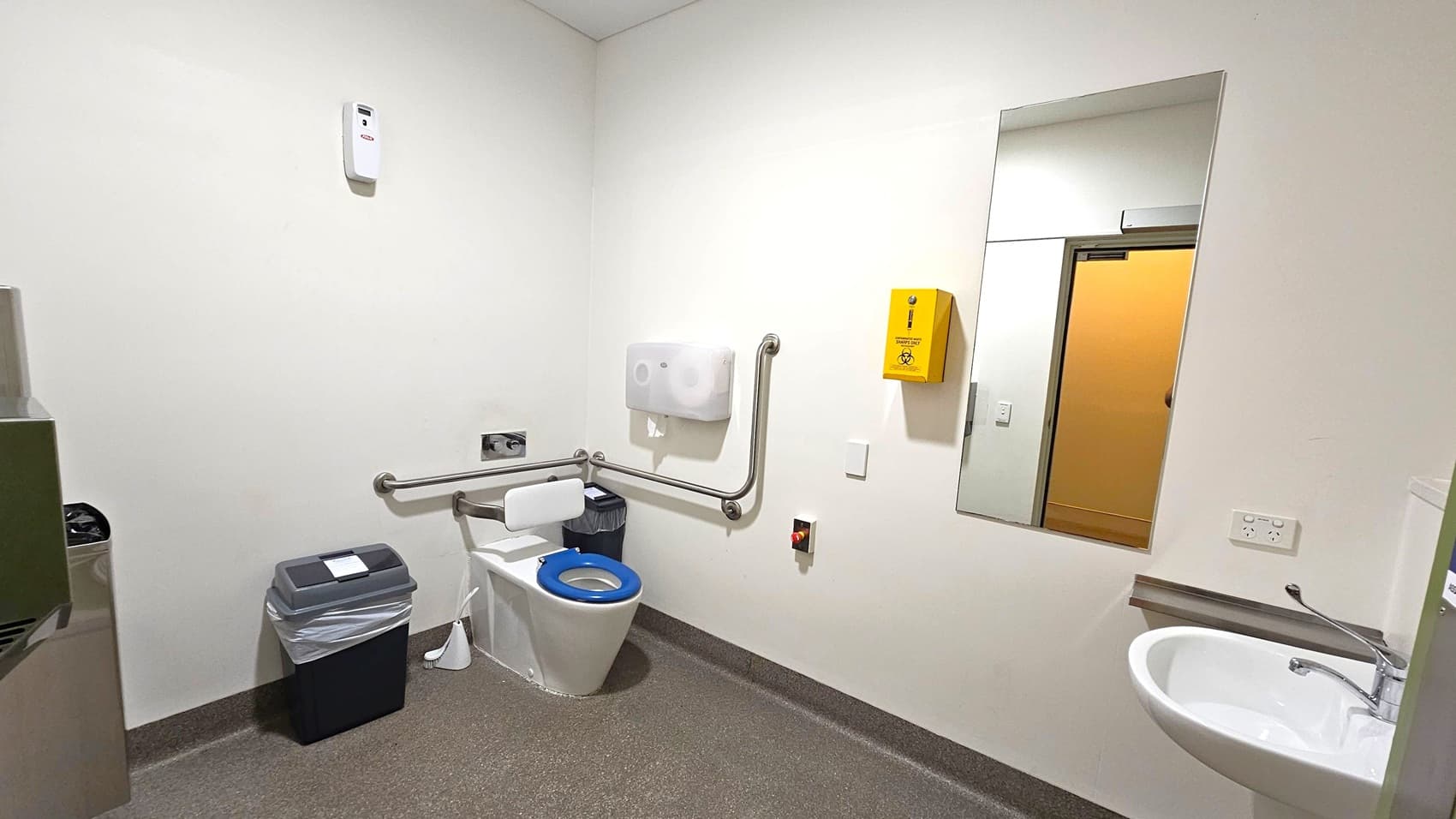 A clean, accessible bathroom with a white toilet featuring a blue seat and support rails on the left. A bin with a liner is next to the toilet. Above, a paper dispenser and a yellow sharps disposal box are mounted on the wall. On the right, a large mirror reflects the opposite wall and a door. Below the mirror, a white sink with a lever tap is installed. The floor is grey with a textured surface, and the walls are plain white.