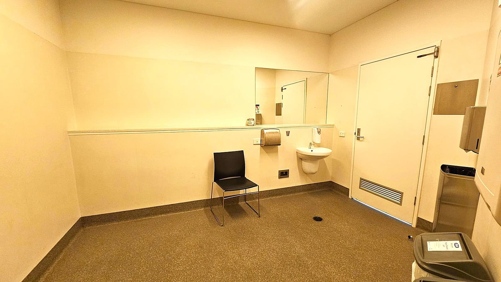 A spacious, beige-walled room with a speckled grey floor. On the right, a white door with a vent at the bottom. Next to it, a small white sink with a soap dispenser above and a paper towel dispenser to the left. A black chair is positioned against the wall beneath a large mirror. In the foreground, a grey bin with a lid. The ceiling is plain with recessed lighting.
