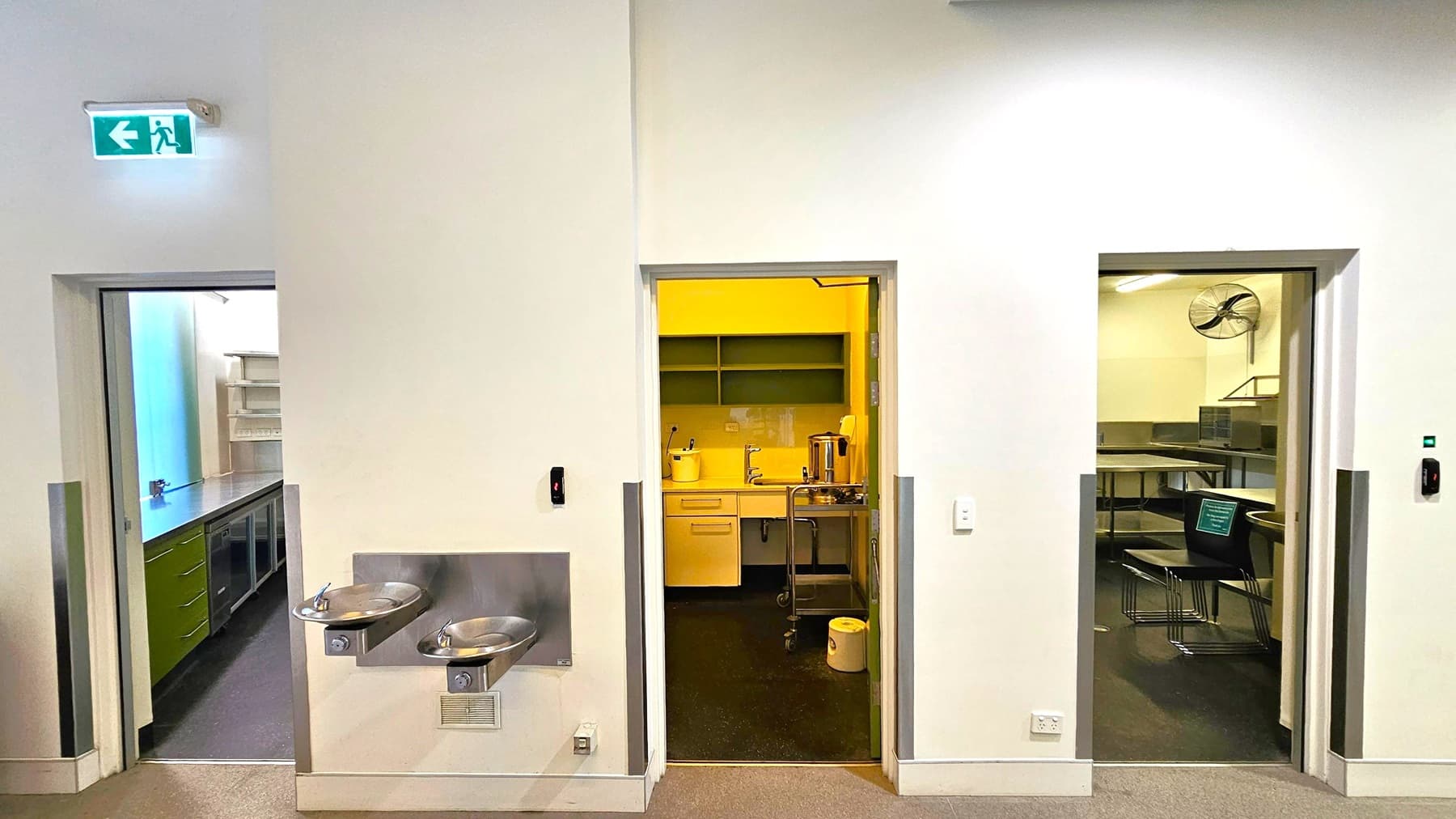 A wall with three doorways. The left doorway leads to a room with shelves and a countertop. Above it, an illuminated green exit sign with a left arrow. In the centre, a doorway opens to a kitchenette with a sink, shelves, and a kettle. Below this doorway, two stainless steel drinking fountains are mounted on the wall. The right doorway reveals a room with tables, chairs, and a wall-mounted fan. The floor is carpeted, and the walls are light-coloured.