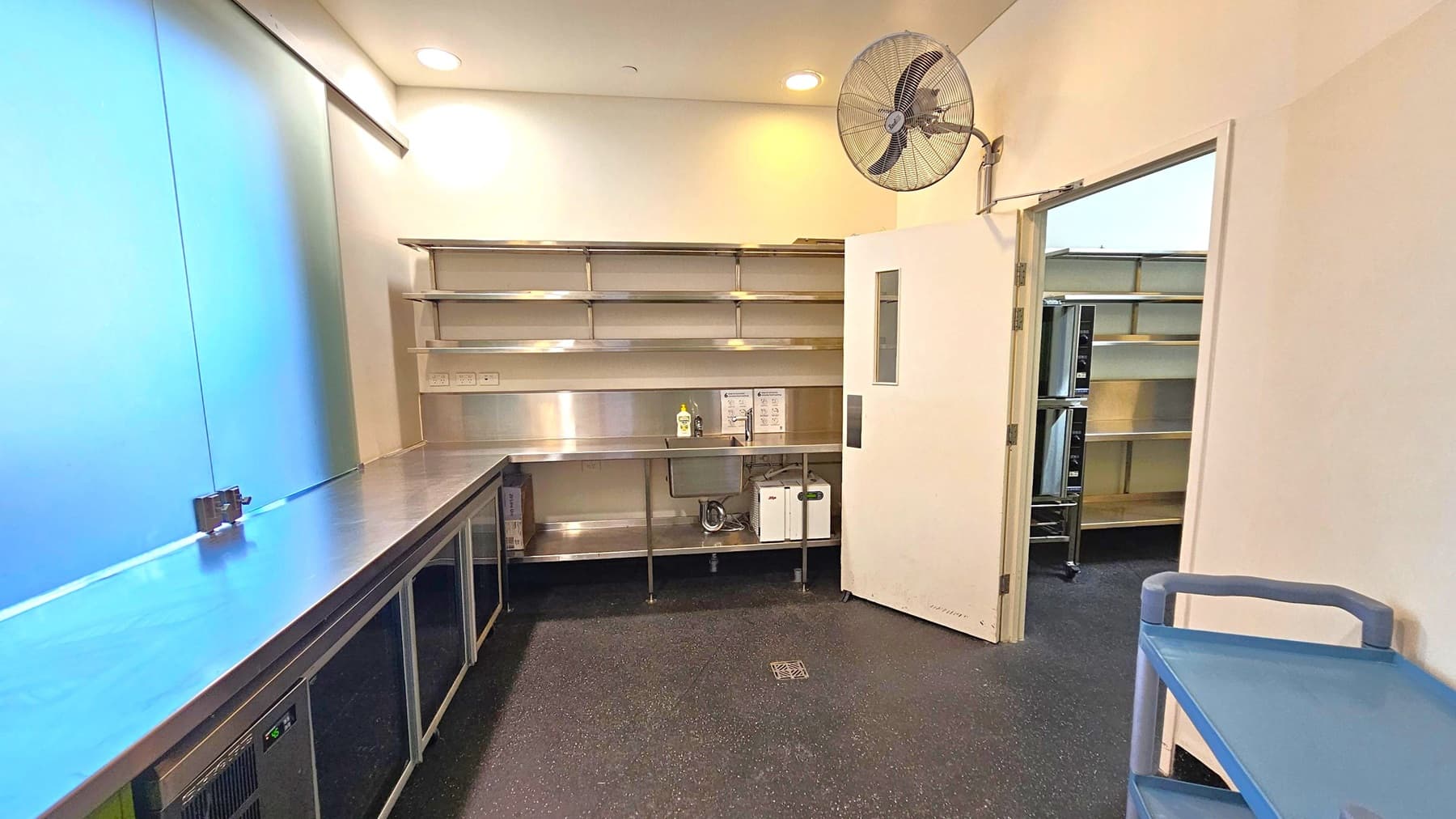 A commercial kitchen with stainless steel countertops and shelving on the left. Below the counter are refrigeration units. On the right, a door is partially open, revealing more shelving in the adjacent room. A wall-mounted fan is above the door. The floor is dark with a speckled pattern, and the ceiling is white with recessed lighting. A blue trolley is in the foreground on the right.