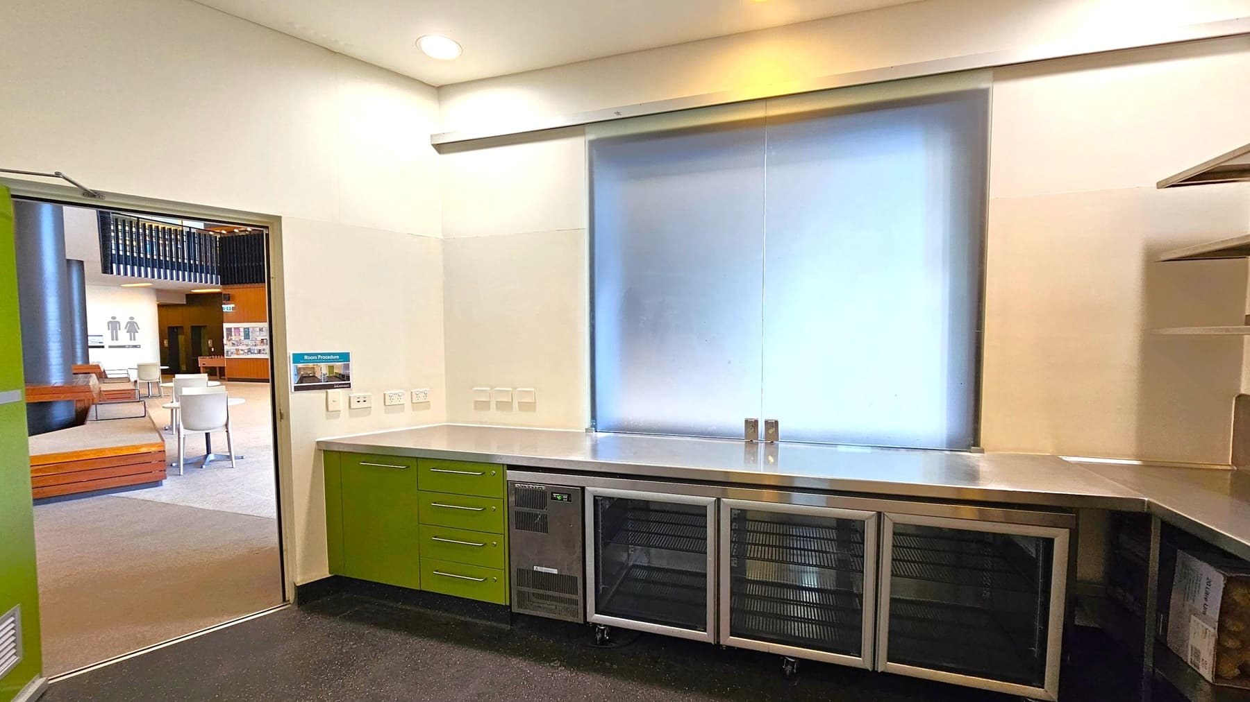 A room with a stainless steel countertop and green cabinets below. There are multiple power outlets on the wall above the counter. A large frosted window is centred on the wall. To the left, an open door leads to a lobby area with chairs and tables. Signs for restrooms are visible in the background. The floor is dark and speckled, and the ceiling is light with recessed lighting. Shelving is partially visible on the right.
