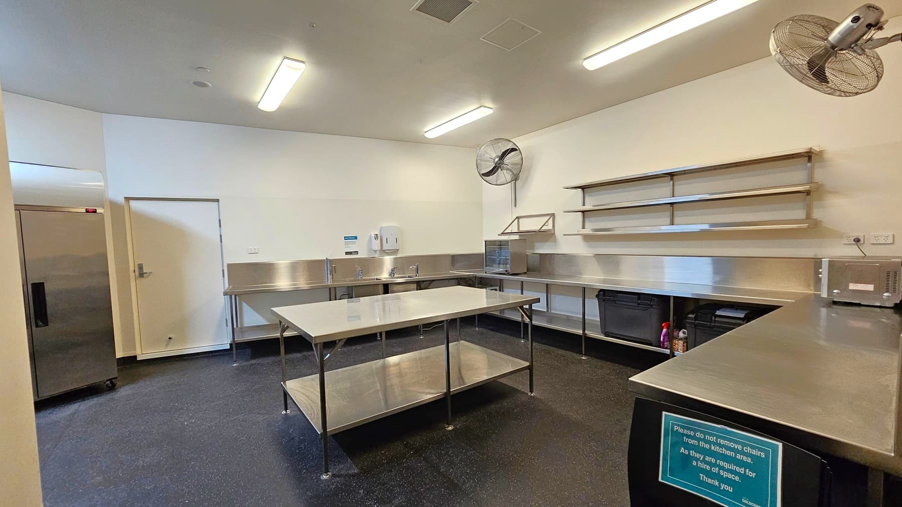 A commercial kitchen with stainless steel worktops and shelving on the right wall. In the centre, there is a large stainless steel table with a lower shelf. On the left, a closed door and a large refrigerator. The floor is dark and speckled, while the ceiling is white with fluorescent lights and wall-mounted fans. A sign on the front worktop advises not to remove chairs from the kitchen area.