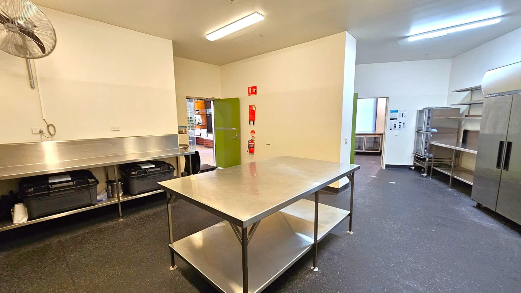 A commercial kitchen with a stainless steel worktable in the centre. On the left, a wall-mounted fan and a long counter with storage underneath. In the background, a green door leads to another room, with fire safety equipment mounted nearby. On the right, stainless steel shelves and a large refrigerator. The floor is dark and speckled, and the ceiling has fluorescent lighting.