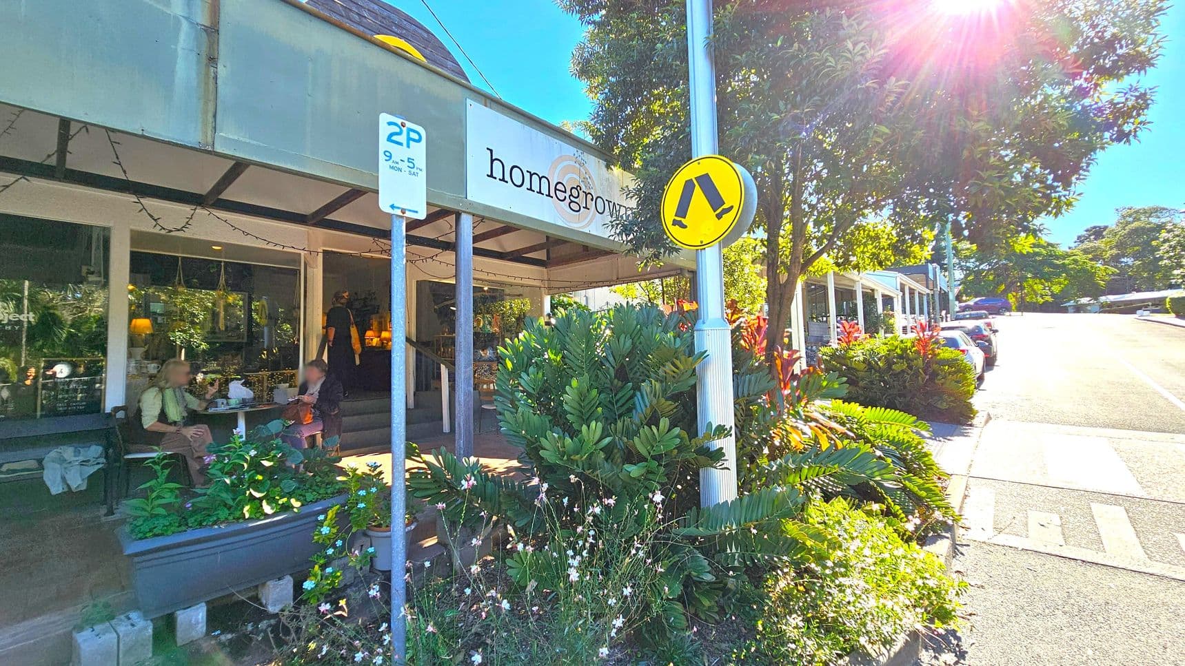 A café with a sign reading "homegrown" on the left, featuring outdoor seating where two people are sitting at a table. A parking sign and a pedestrian crossing sign are visible near lush green plants. On the right, a street lined with parked cars extends uphill, bordered by trees and more greenery. Sunlight filters through the trees, casting shadows on the pavement.