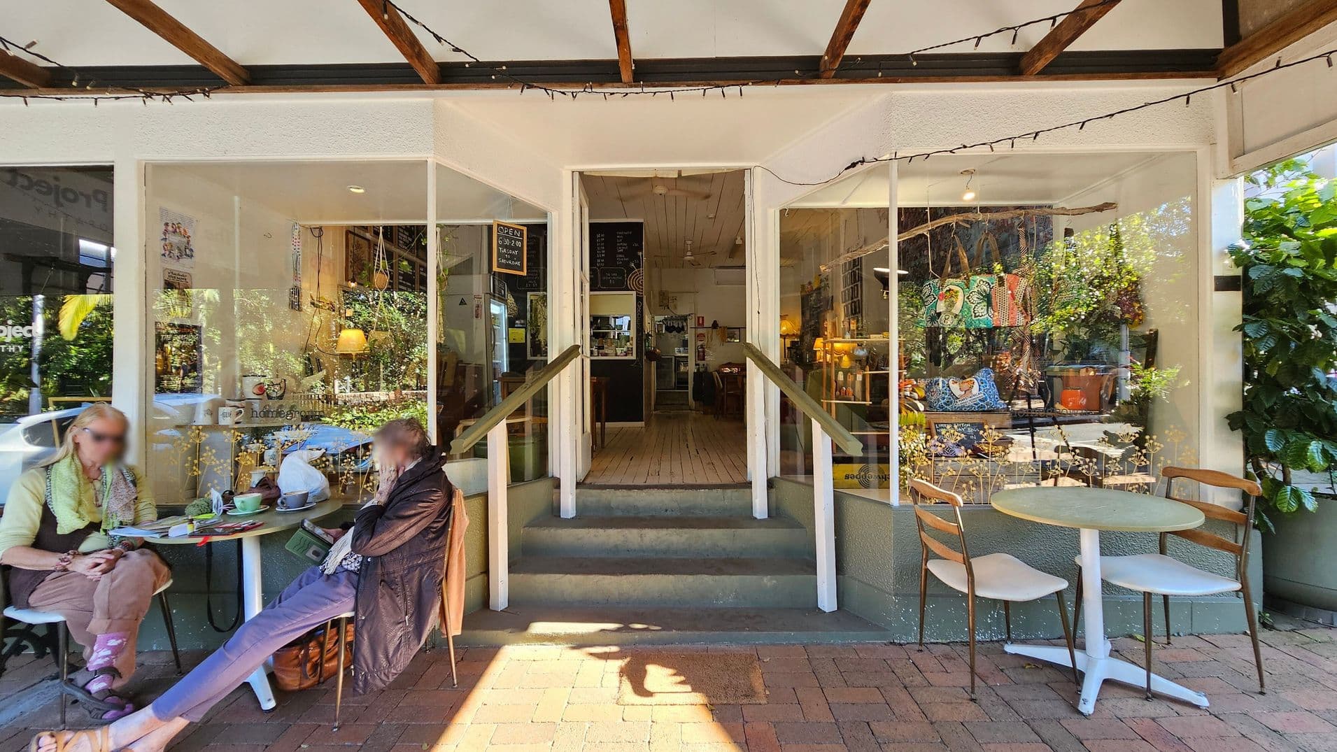A small café entrance with two large windows on either side. On the left, two people sit at a round table with drinks and papers. On the right, another round table with two empty chairs. Inside, a wooden floor leads to a counter with a menu board above. The windows display colourful bags and plants. The ceiling has exposed wooden beams with string lights, and the floor is brick.