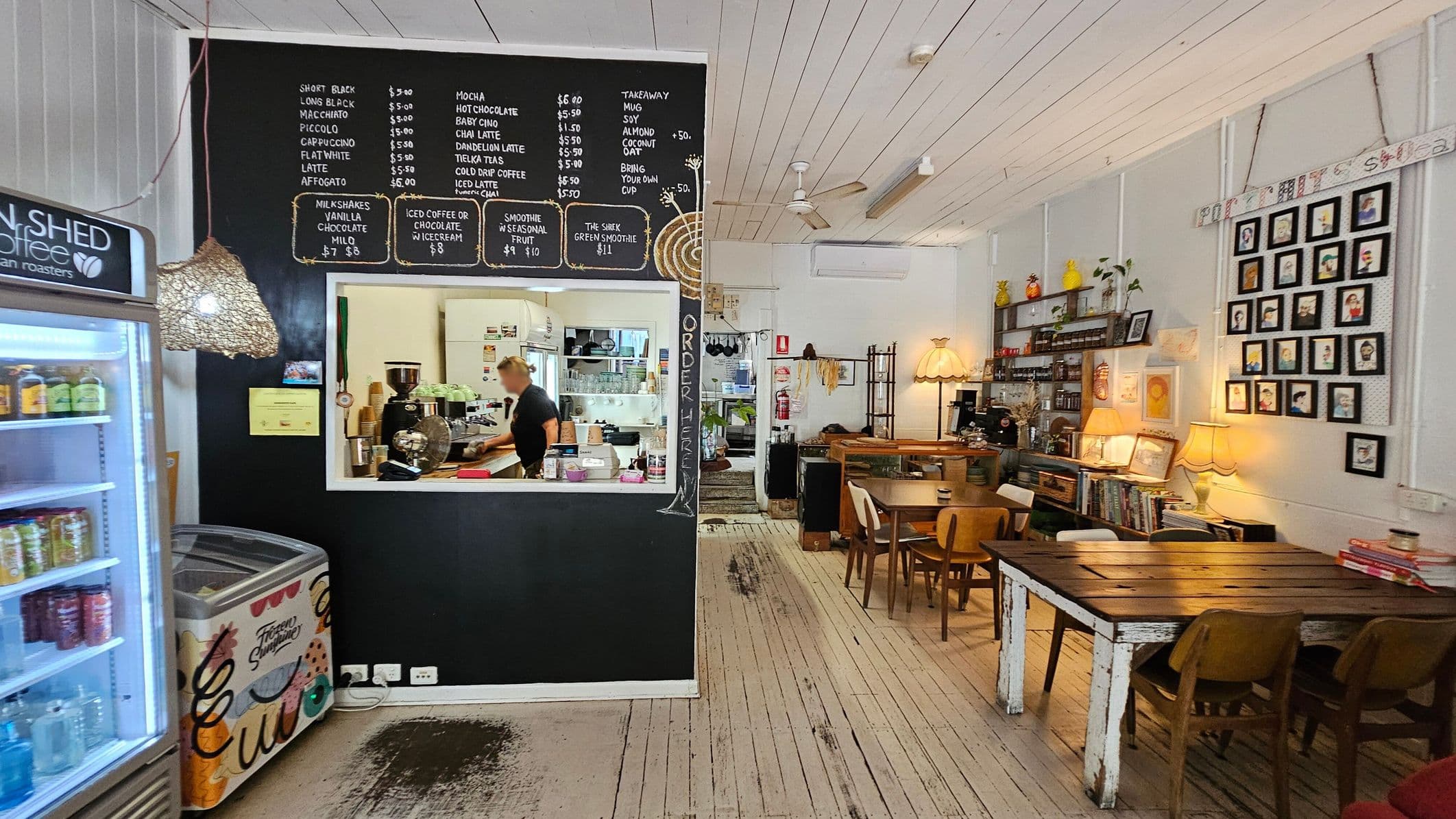 A cosy café interior with a blackboard menu on the left wall above a serving window, where a person is preparing drinks. A fridge with drinks and an ice cream freezer are nearby. On the right, wooden tables and chairs are arranged, with a wall displaying framed pictures. Shelves hold various items, and warm lighting from lamps creates a welcoming atmosphere. The floor is wooden, and the ceiling is white with exposed beams and a ceiling fan.