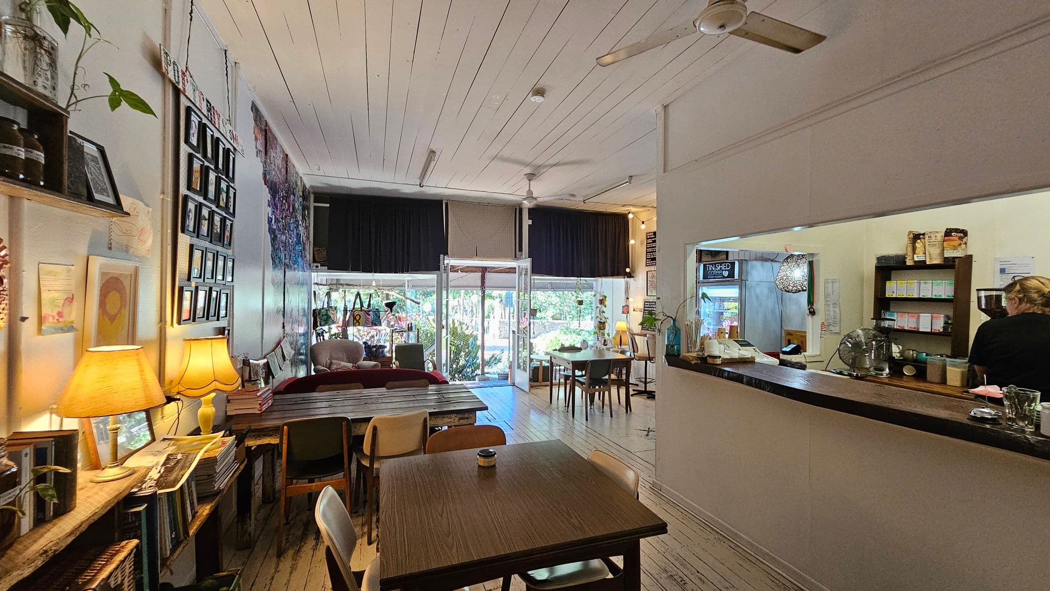 A cosy café interior with wooden flooring and a white panelled ceiling. On the left, a wall displays framed photos and artwork, with a shelf holding books and a lamp. In the centre, tables and chairs are arranged, leading to a glass entrance with views of greenery outside. On the right, a counter with a person working behind it, shelves stocked with products, and a hanging light fixture. Ceiling fans are visible above.