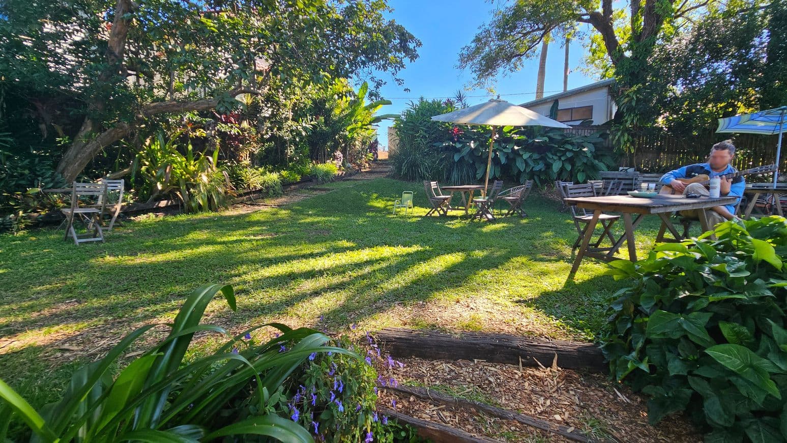 A garden with a grassy area surrounded by lush greenery and trees. On the left, there are two wooden chairs near dense foliage. In the centre, a pathway leads through the garden, with a table and chairs under a large umbrella. On the right, a person is seated at a wooden table playing a guitar. The foreground has vibrant plants and flowers, while the background shows more trees and a wooden fence. Shadows from the trees stretch across the grass.