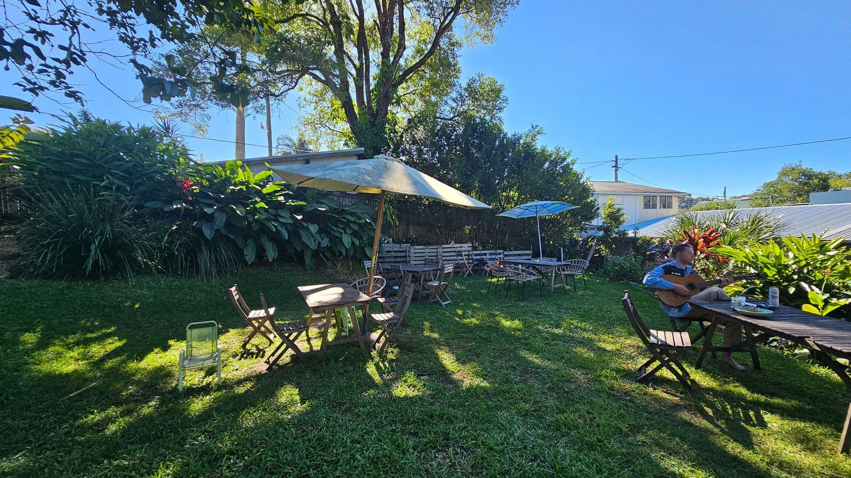 A garden setting with lush greenery and large plants in the background. Several wooden tables and chairs are scattered across the grass, with some tables shaded by large umbrellas. On the right, a person is seated at a table playing a guitar. A house with a sloped roof is visible in the background, partially obscured by foliage. The scene is well-lit, suggesting a sunny day.