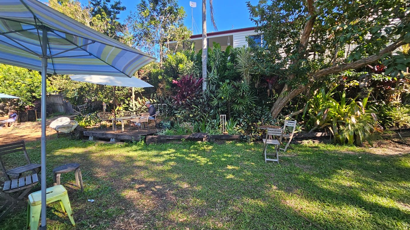 A garden setting with a grassy area in the foreground. On the left, a large striped umbrella shades a wooden chair and a small stool. In the centre, a wooden deck with a table and chairs is partially shaded by another umbrella. The background is filled with lush greenery, including various plants and trees. A house is partially visible behind the foliage. A few more chairs are scattered around the garden.