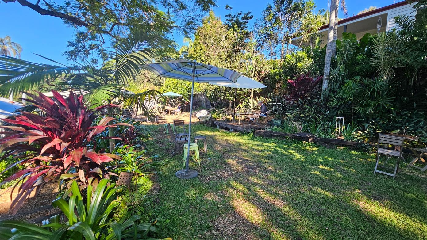 A garden with lush greenery and various plants, including red and green foliage on the left. In the centre, there are several outdoor tables and chairs, some with umbrellas providing shade. The ground is covered with grass, and there are patches of sunlight and shadow. In the background, a house is partially visible, surrounded by tall trees and dense vegetation.
