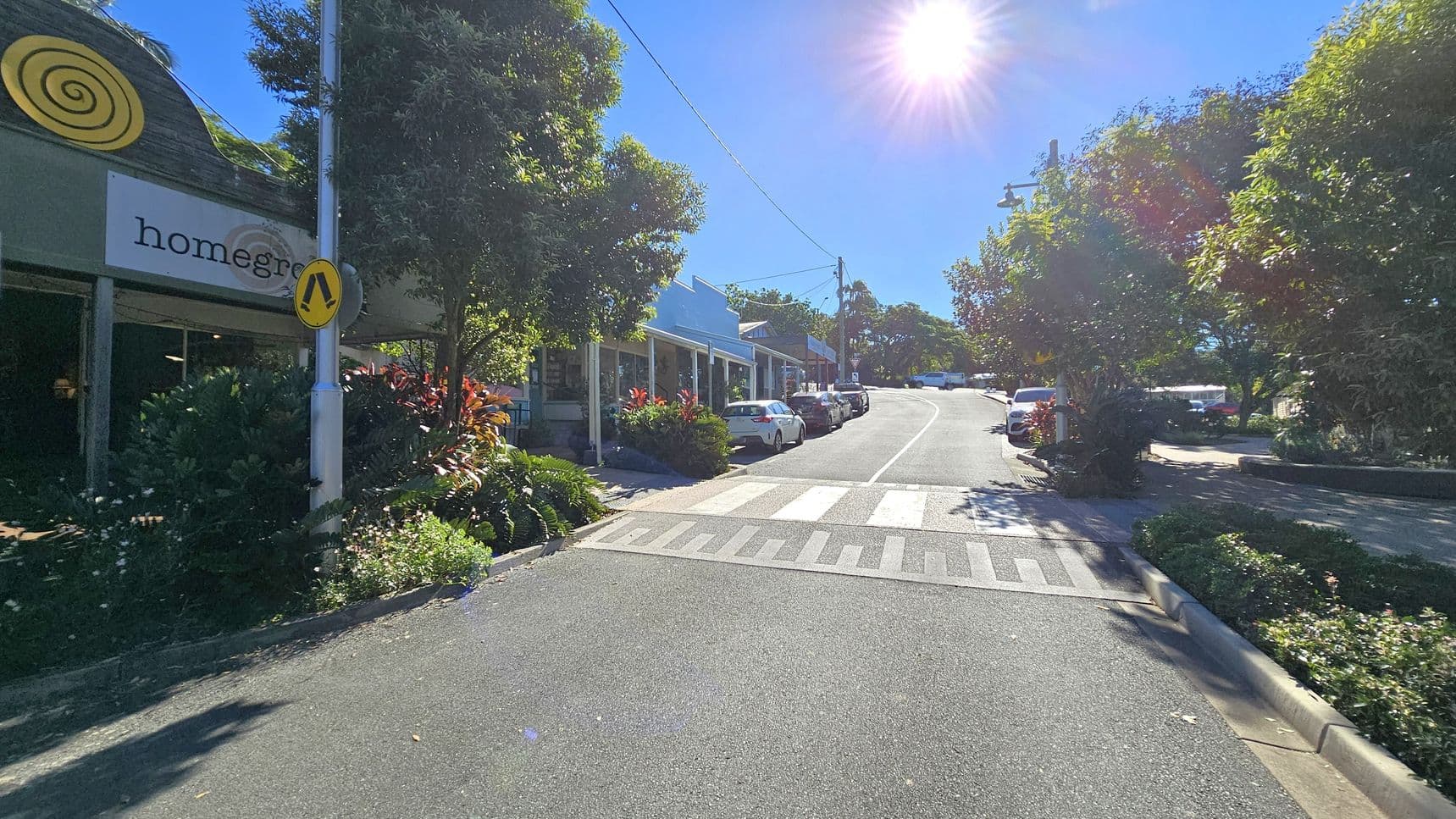 A street scene with a pedestrian crossing in the foreground. On the left, a building with a green exterior and a sign reading "homegrown" is partially visible, surrounded by lush greenery. On the right, a row of parked cars lines the street, which curves uphill. Trees and bushes are abundant on both sides, and the sun is shining brightly in the clear blue sky, casting shadows on the road.