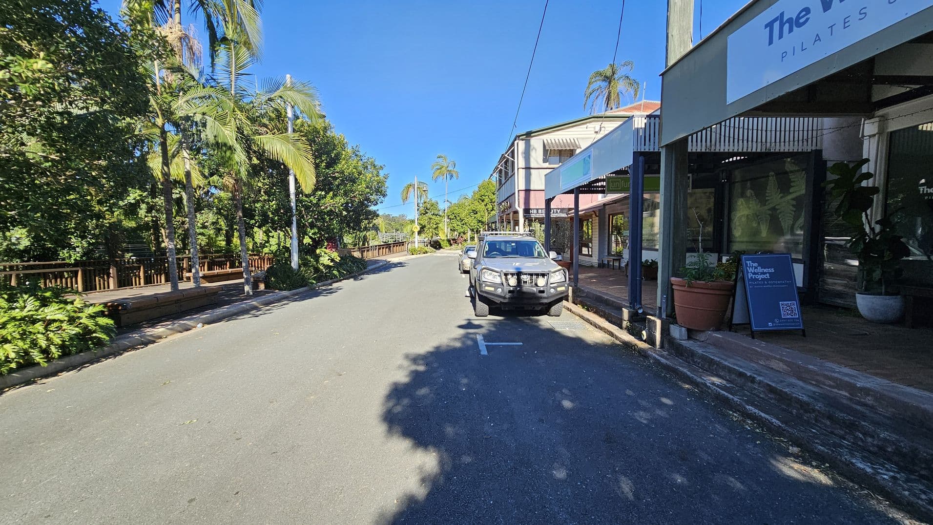 A street scene with a vehicle parked on the right side near a row of shops. The shop signs include "The Wellness Project" and "HB Beauty." Large potted plants are placed outside the shops. On the left, there are tall palm trees and dense greenery along a wooden fence. The road is clear, with shadows from the trees cast on the pavement. The sky is clear and blue.