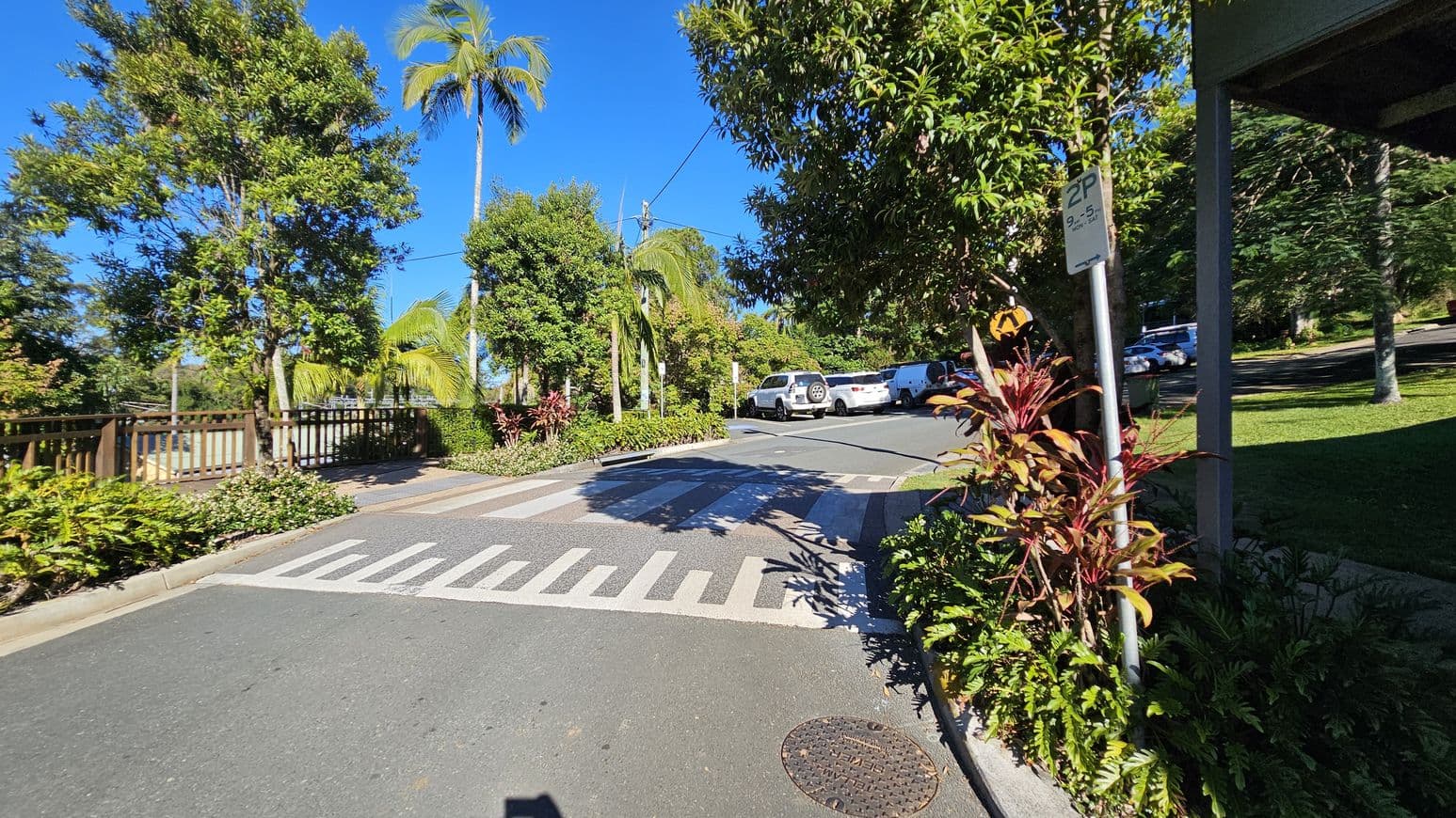 A street scene with a pedestrian crossing in the foreground. On the right, a signpost with parking information is surrounded by red and green foliage. Several cars are parked in a lot further back. On the left, a wooden fence runs alongside the road, with tall palm trees and lush greenery. The road surface is smooth, and the sky is clear and blue.