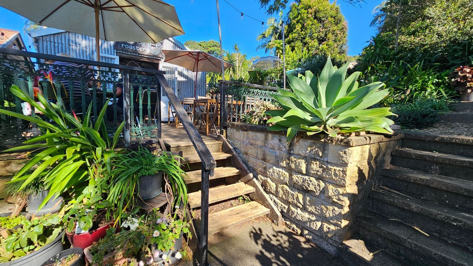 A small outdoor café area with wooden steps leading up to a deck on the left. The deck is bordered by a decorative metal railing and shaded by large umbrellas. Several potted plants, including a large succulent, are placed around the area. To the right, stone steps ascend alongside a stone wall, surrounded by lush greenery. The background features a corrugated metal building with a sign partially visible. The ground is paved, and string lights are strung above.