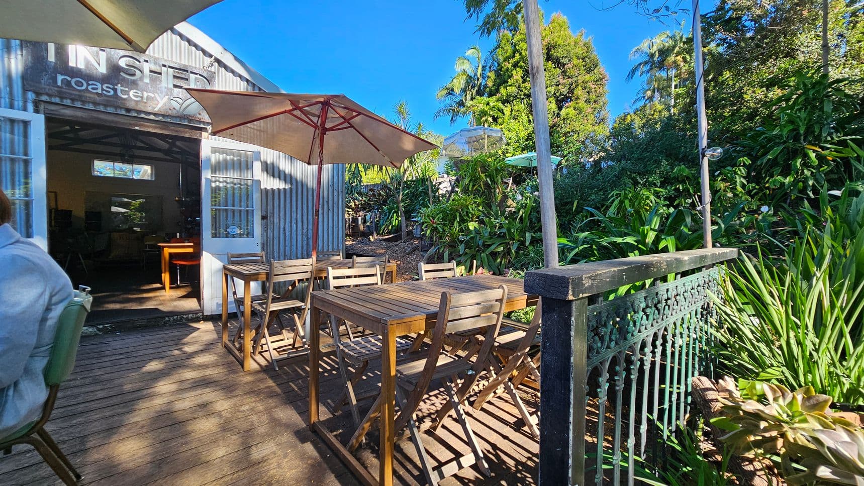 Outdoor seating area with wooden tables and chairs under large umbrellas. On the left, a corrugated metal building with an open door and a sign reading "roastery." Lush greenery and various plants surround the area, with a decorative metal railing on the right. The floor is wooden decking, and the sky is clear and blue.