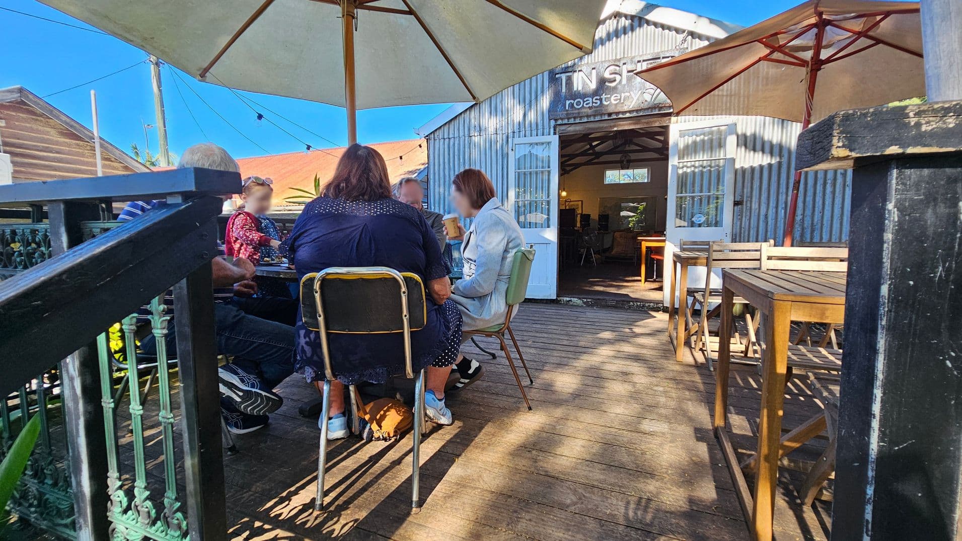 A group of people sit around a table under a large umbrella on a wooden deck. To the right, there are empty wooden tables and chairs. In the background, a corrugated metal building with an open door displays the words "Tin Shed Roastery." The deck is bordered by a decorative metal railing on the left. The scene is well-lit, suggesting a sunny day.