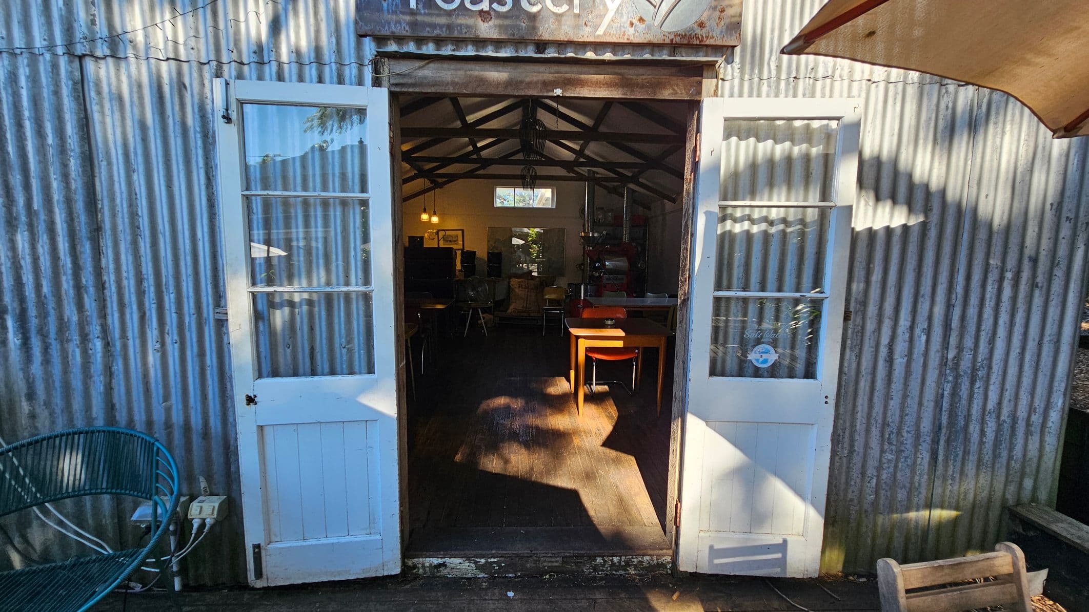 Corrugated metal building with open double doors leading inside. The doors are white with glass panes. Inside, a wooden table and chairs are visible on a wooden floor. The interior has a pitched roof with exposed beams. On the left, a blue metal bench is outside, and on the right, part of a wooden bench and an umbrella are visible. A sign above the doors reads "Roastery."